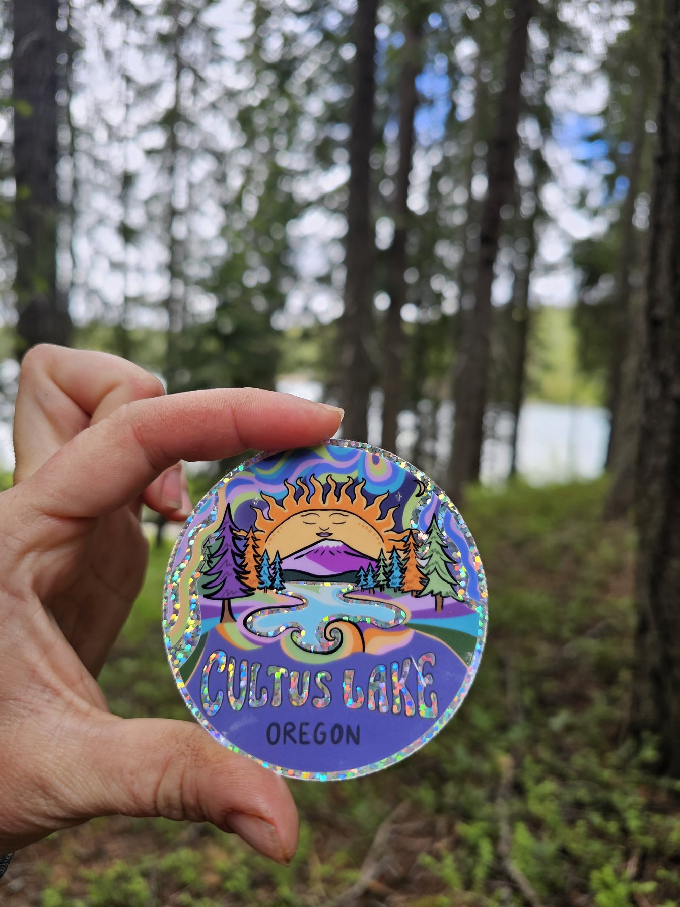 Circular holographic glitter sticker of a sun setting behind a mountain and lake, reading Cultus Lake Oregon