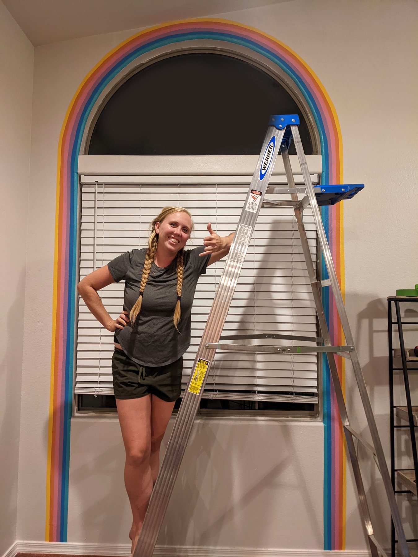 Artist standing beside the finished rainbow arch mural with a ladder